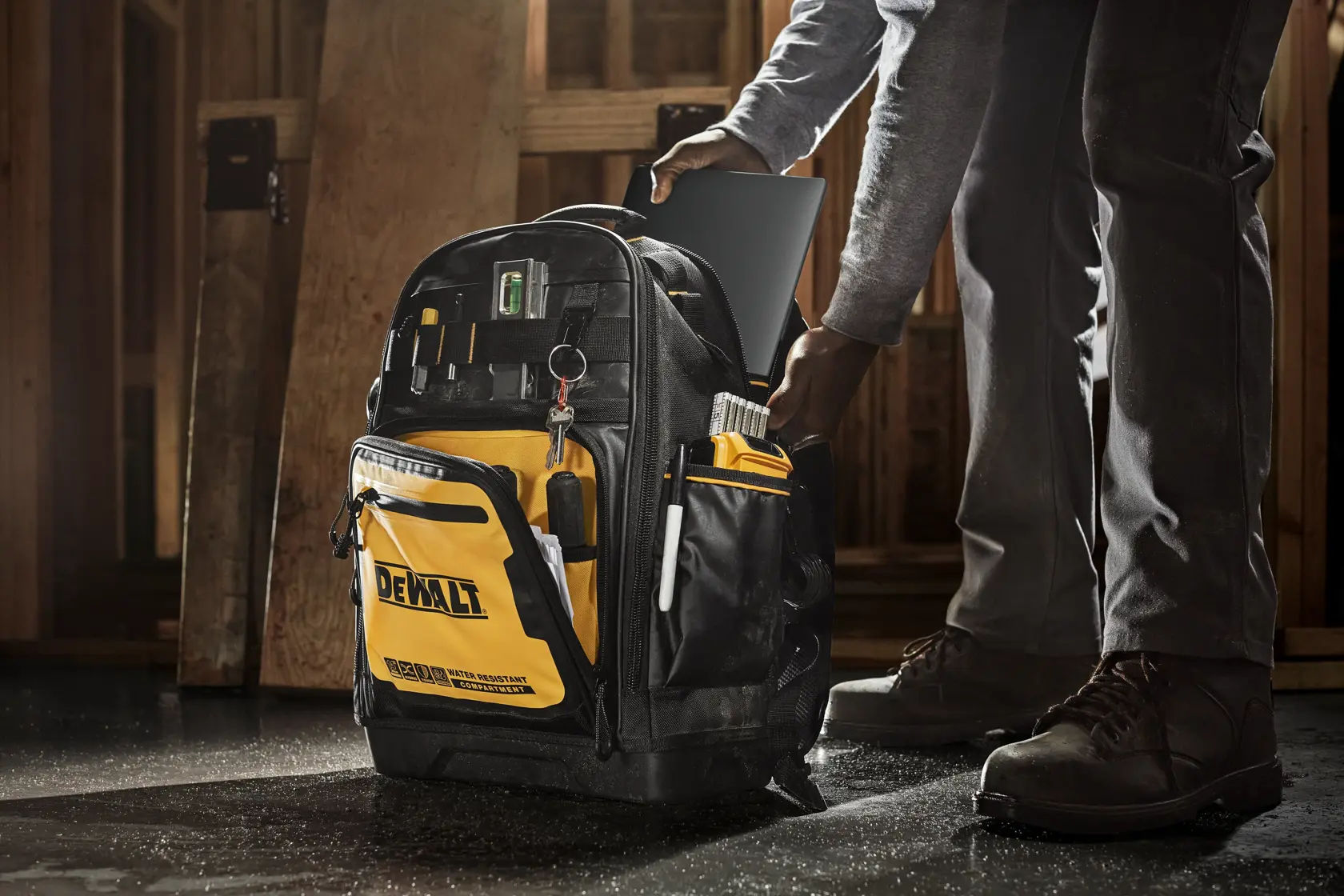 A person places a laptop into a DEWALT tool backpack, which features multiple pockets, organization compartments, and a prominent yellow and black design, standing on a workshop floor.