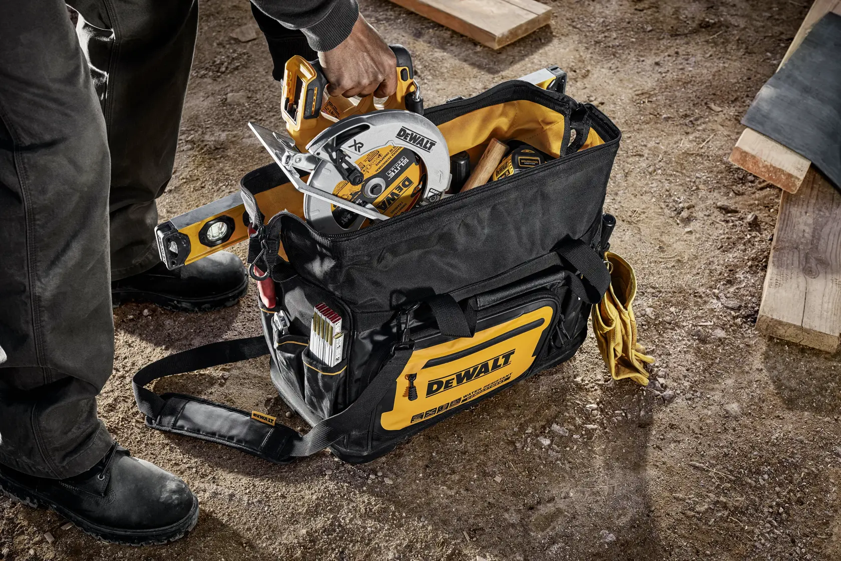 A DEWALT tool bag placed on a construction site floor, filled with various DEWALT tools and accessories including a circular saw, a level, and other hand tools. A person is reaching into the bag to take out the circular saw.