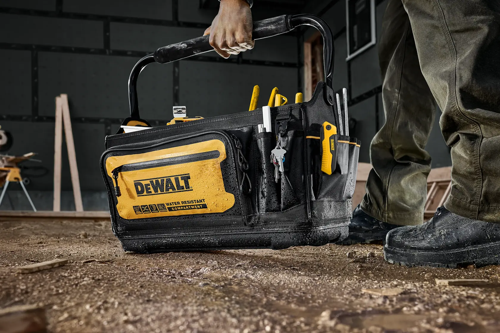 A DEWALT-branded black and yellow tool bag, filled with hand tools, is being carried by a person on a construction site with a dirt floor.