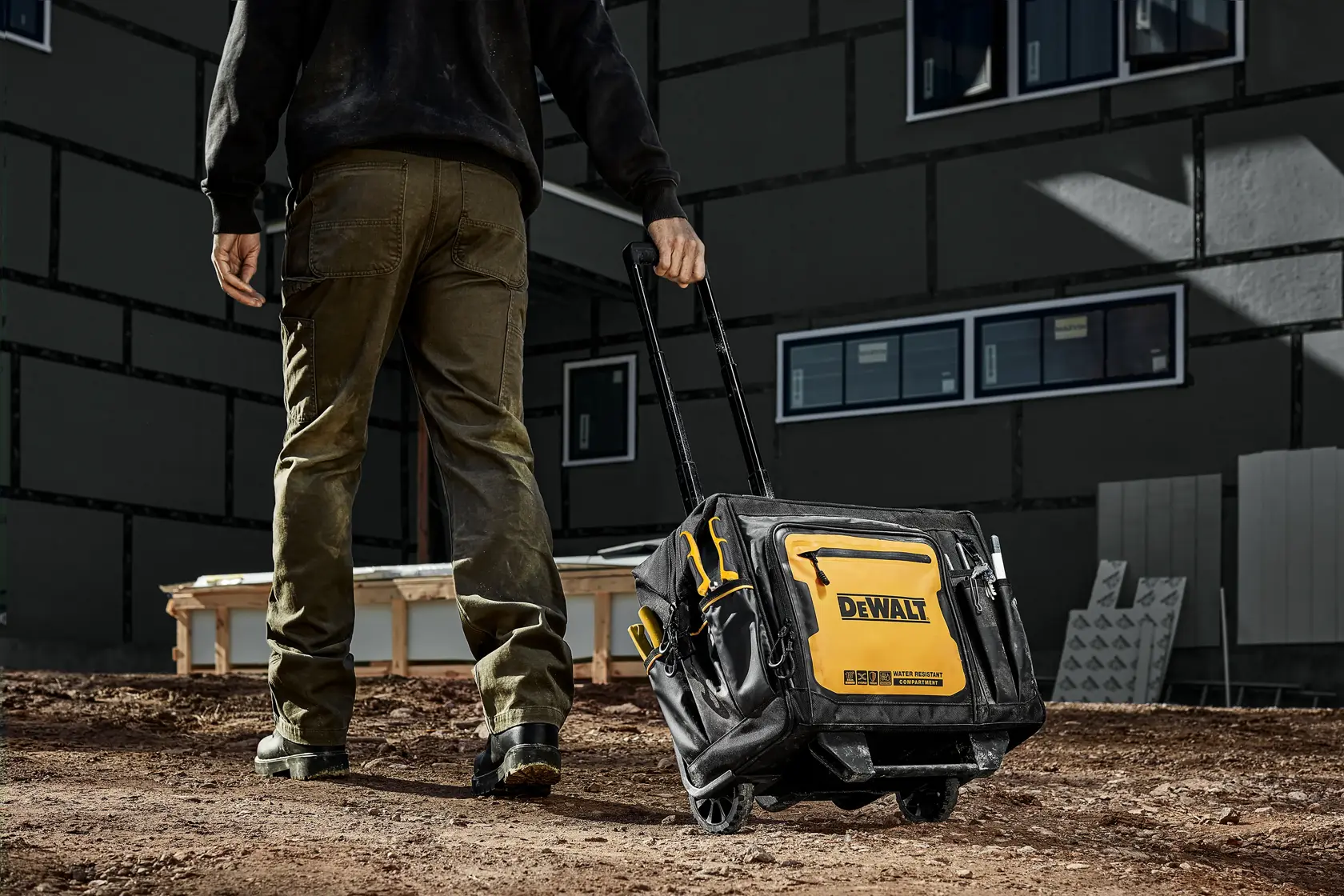 A person is pulling a DEWALT rolling tool bag with a telescopic handle across a dirt construction site. The bag is black and yellow, labeled with the DEWALT logo, and has several compartments for storage.