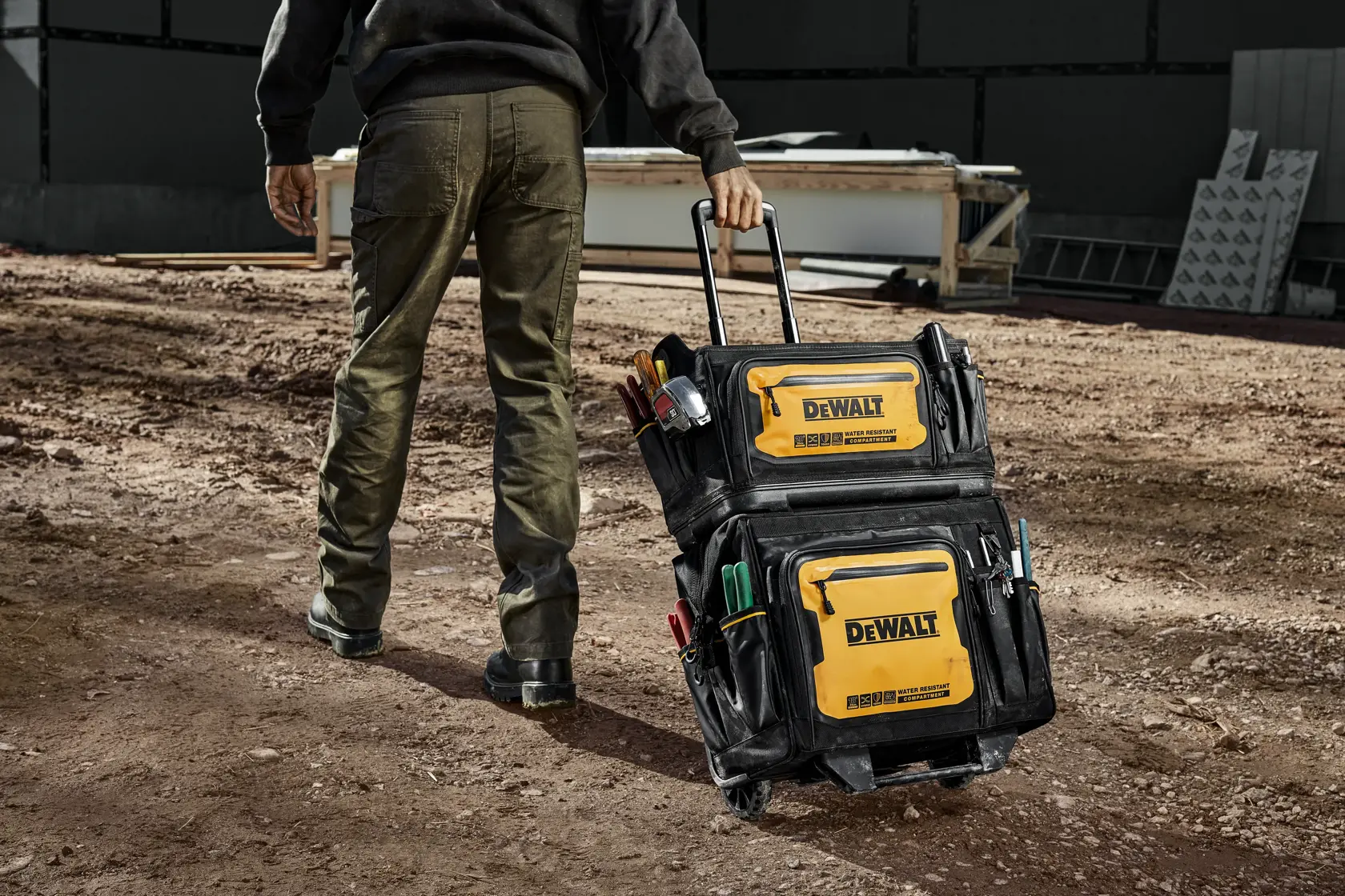 A person is pulling a DEWALT rolling tool bag on a construction site. The bag is black and yellow with multiple pockets and compartments, and is filled with various tools.
