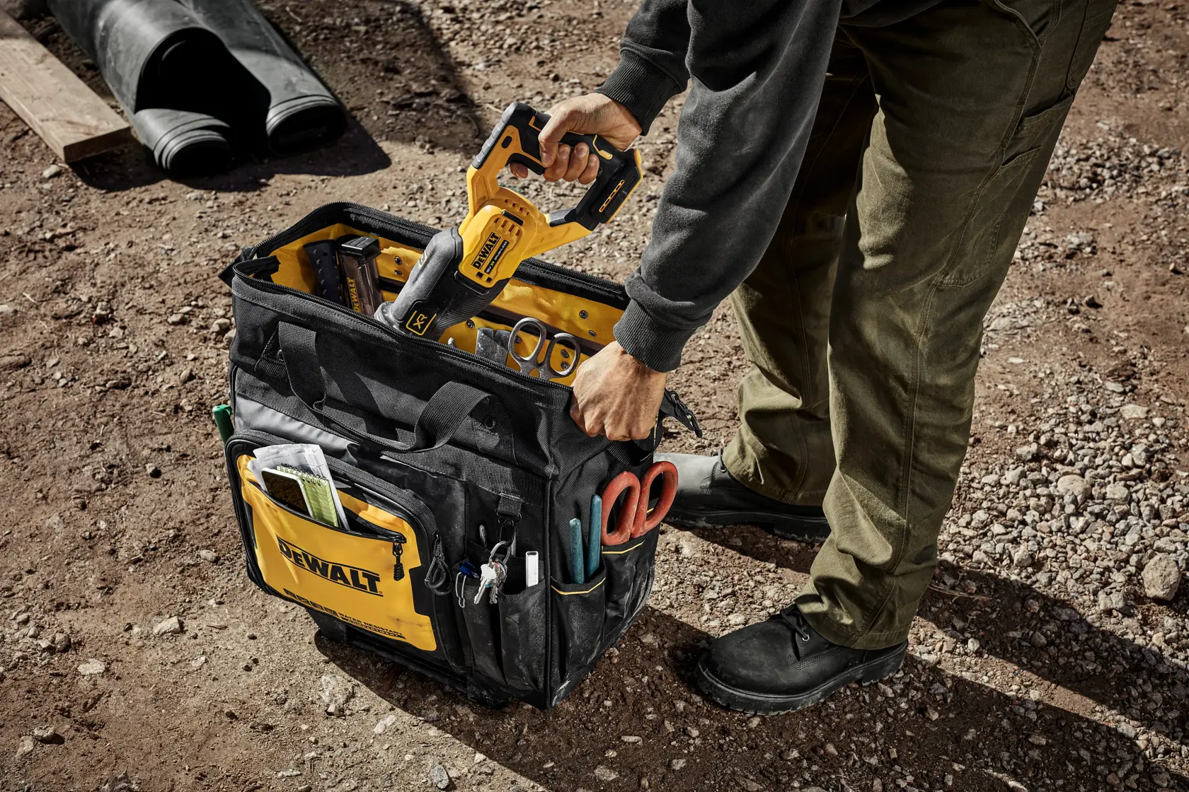 A person placing a yellow DEWALT power tool into a black and yellow DEWALT tool bag filled with various tools and accessories, on a dirt ground at an outdoor work site.