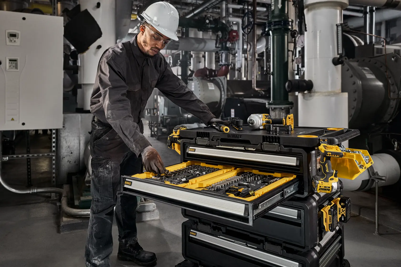 Worker with helmet uses a DEWALT tool storage unit in an industrial setting, accessing organized drawers of tools.