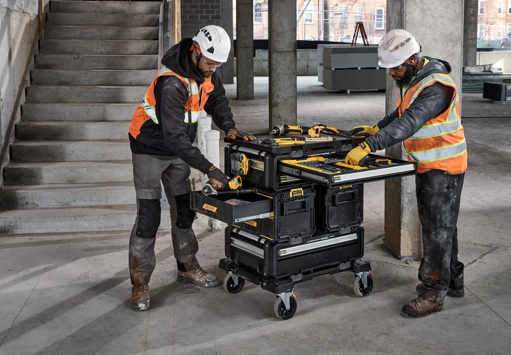 Two workers use a DEWALT rolling tool station with open drawers and tools on top at a construction site.