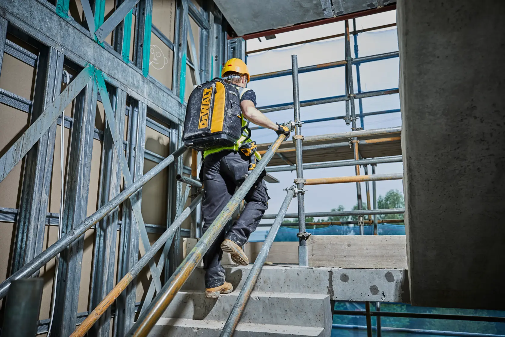 Worker climbs stairs at a construction site carrying a yellow DEWALT backpack.