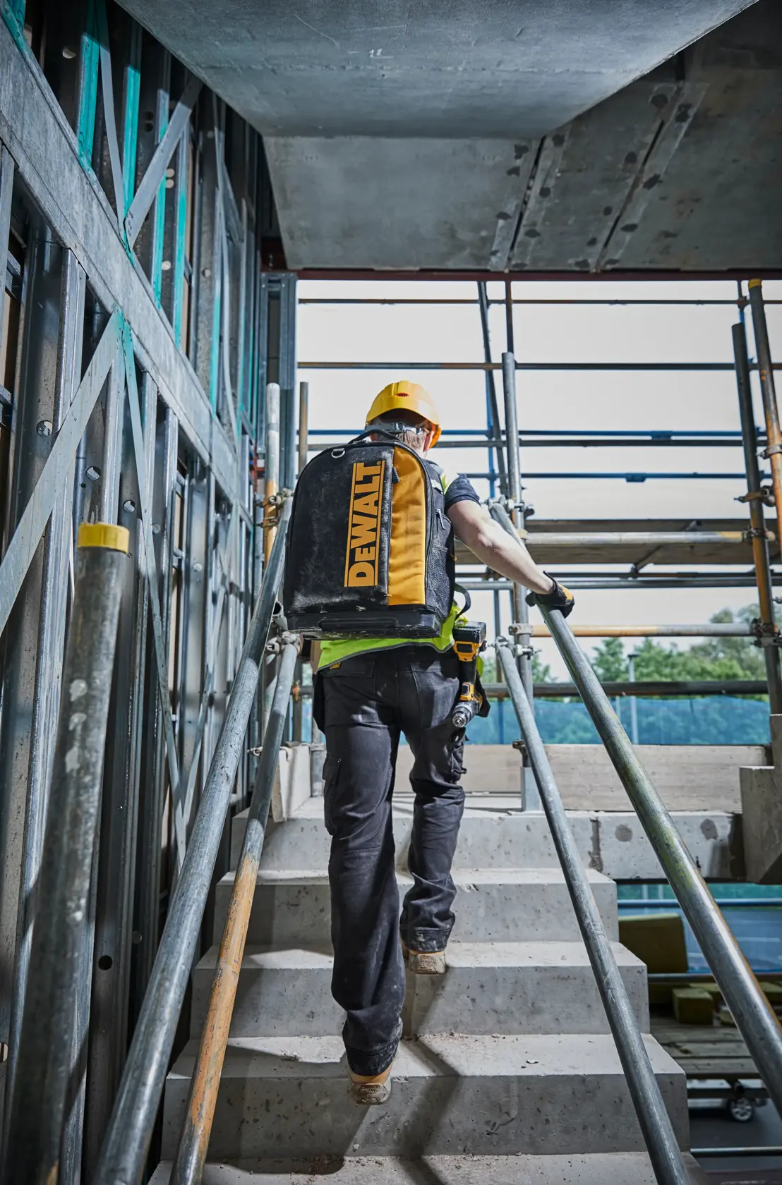 Worker with DEWALT backpack climbing stairs at a construction site, wearing a yellow helmet and safety gear.