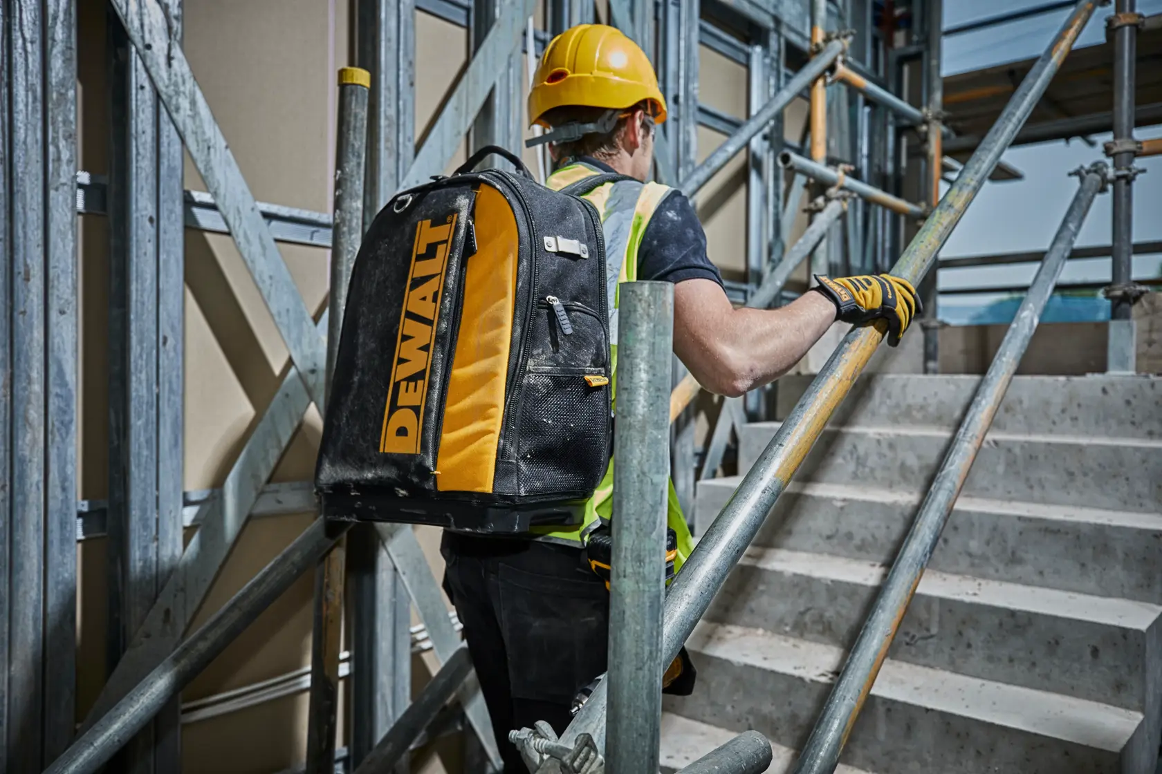 Worker with DEWALT backpack ascending stairs on a construction site, wearing safety gear and helmet.