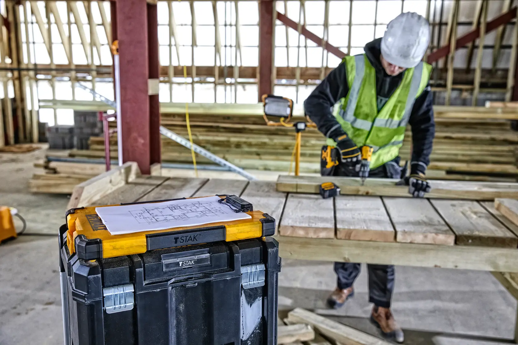 A DEWALT TSTAK toolbox with yellow and black design is placed in the foreground of a construction site. On top of the toolbox is a sheet of paper with diagrams. In the background, a construction worker wearing a hard hat and safety vest is working with a power tool on wooden planks.