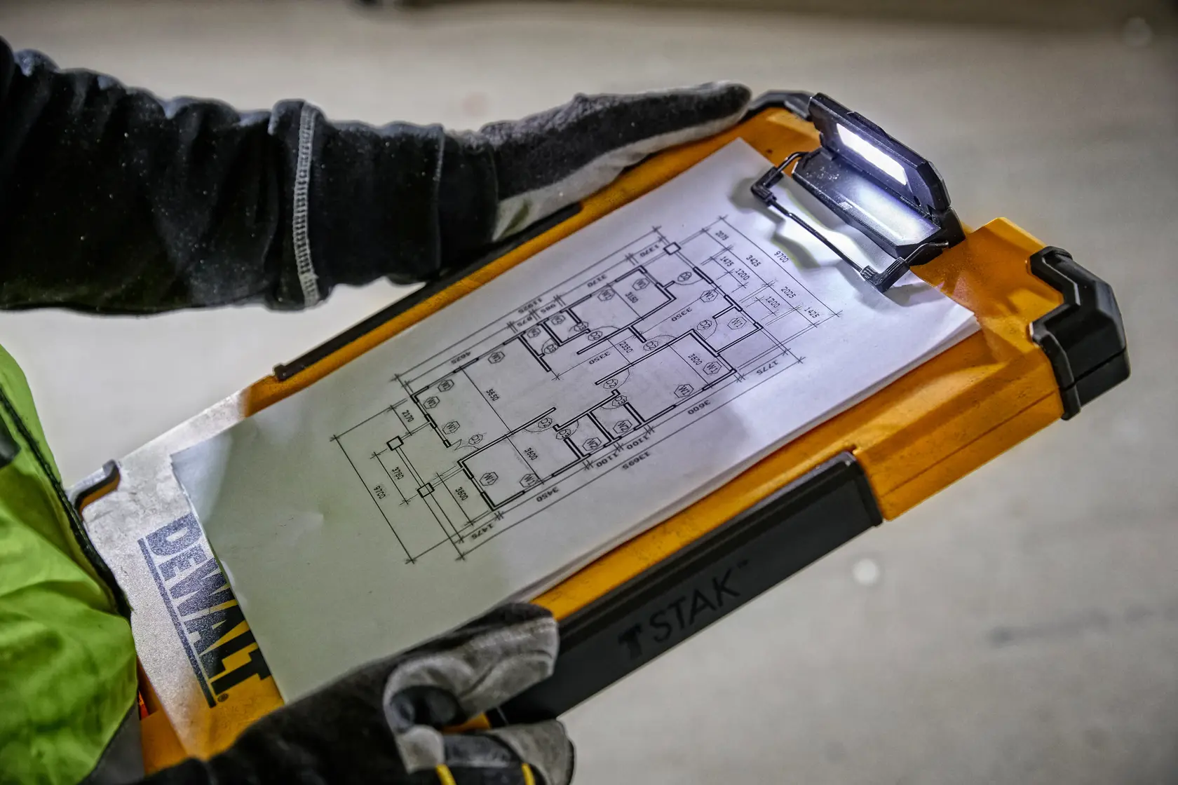 A person wearing gloves holds a DEWALT TSTAK clipboard with an integrated LED light, displaying a technical floor plan drawing attached to the clip.