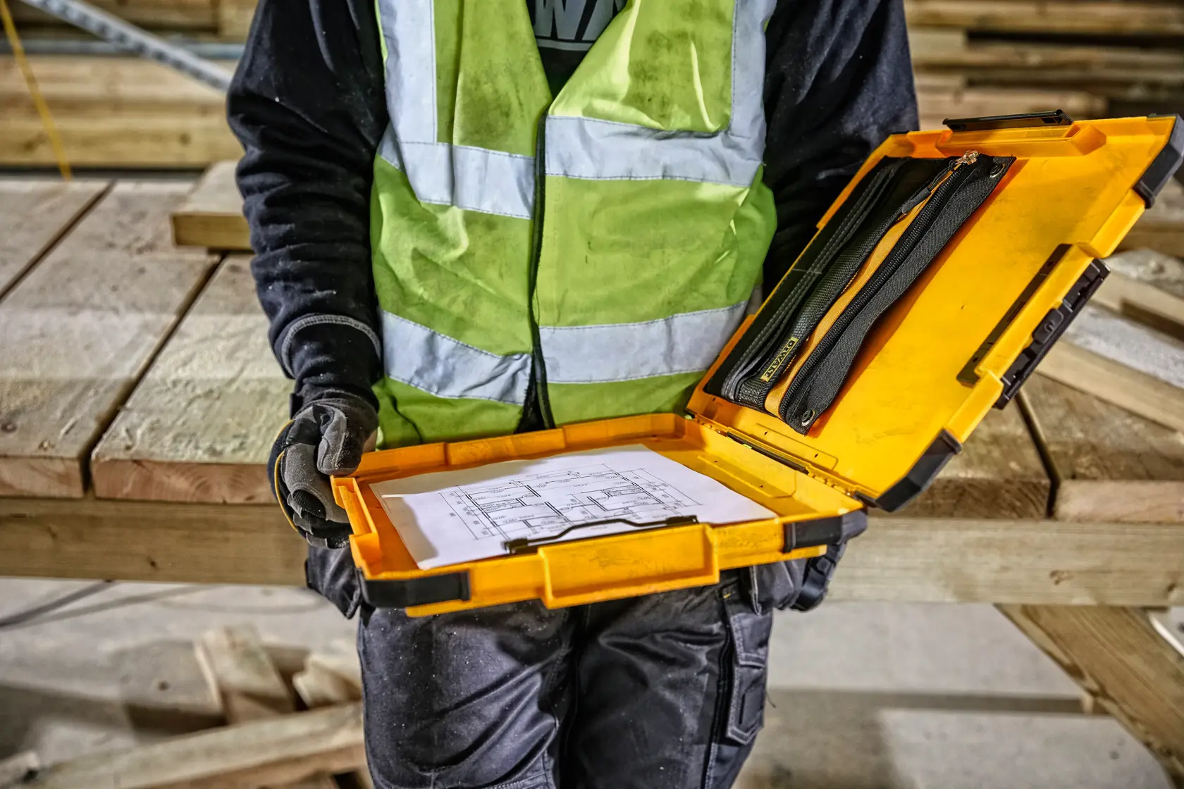 A person wearing a reflective safety vest holds an open yellow DEWALT case containing documents and tools at a construction site with wooden beams in the background.