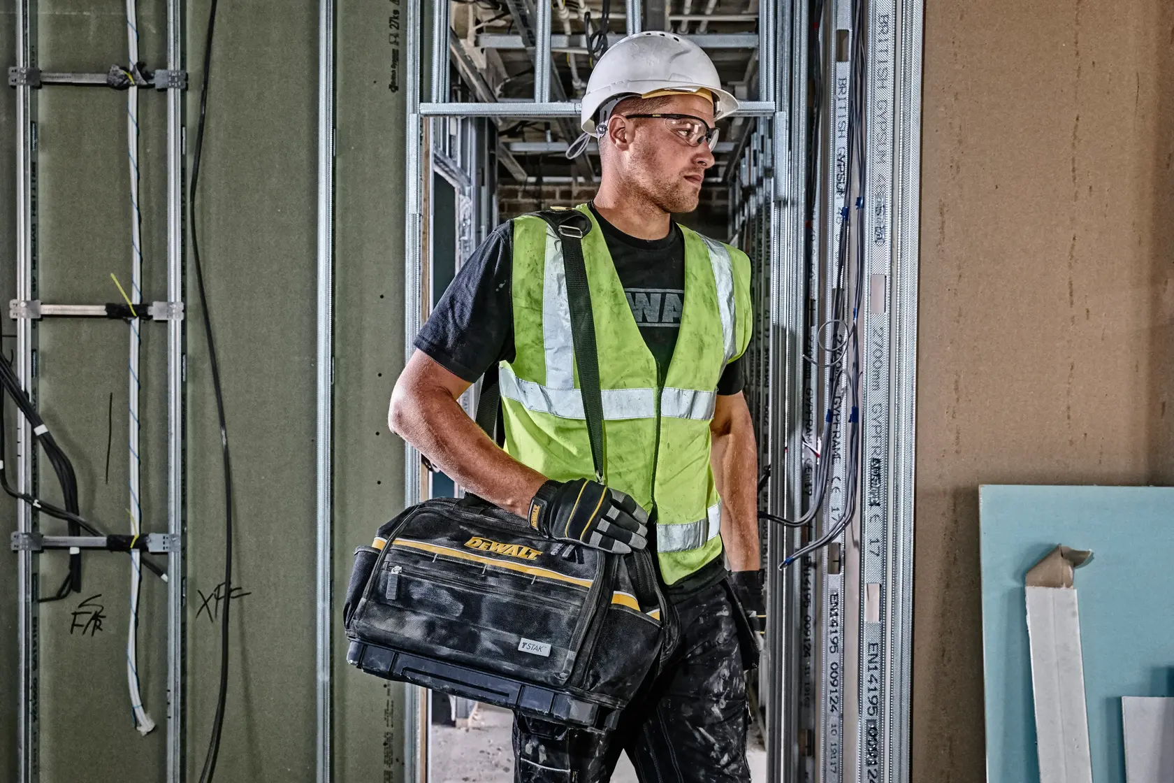 Trabajador con equipo de seguridad y bolso DEWALT en obra con paredes y cableado visibles.