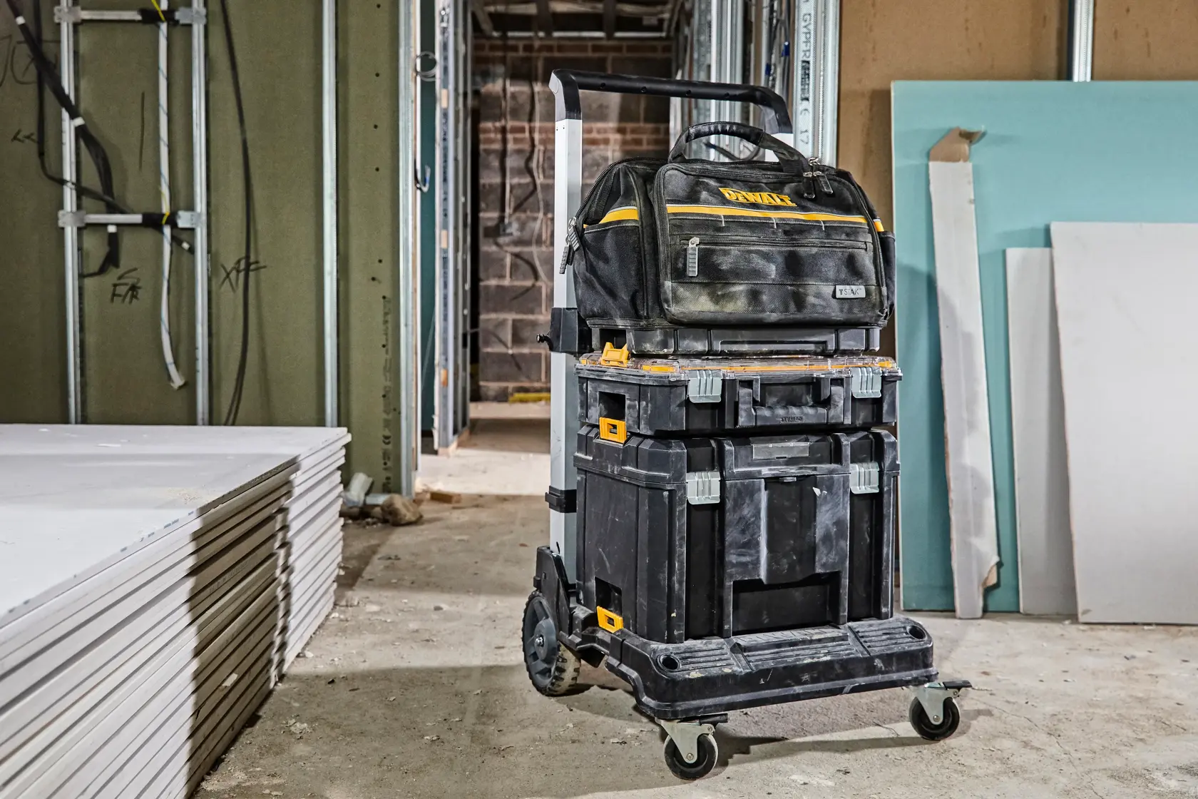 Carro DEWALT con cajas y bolsa apiladas en obra, placas de yeso en el suelo.