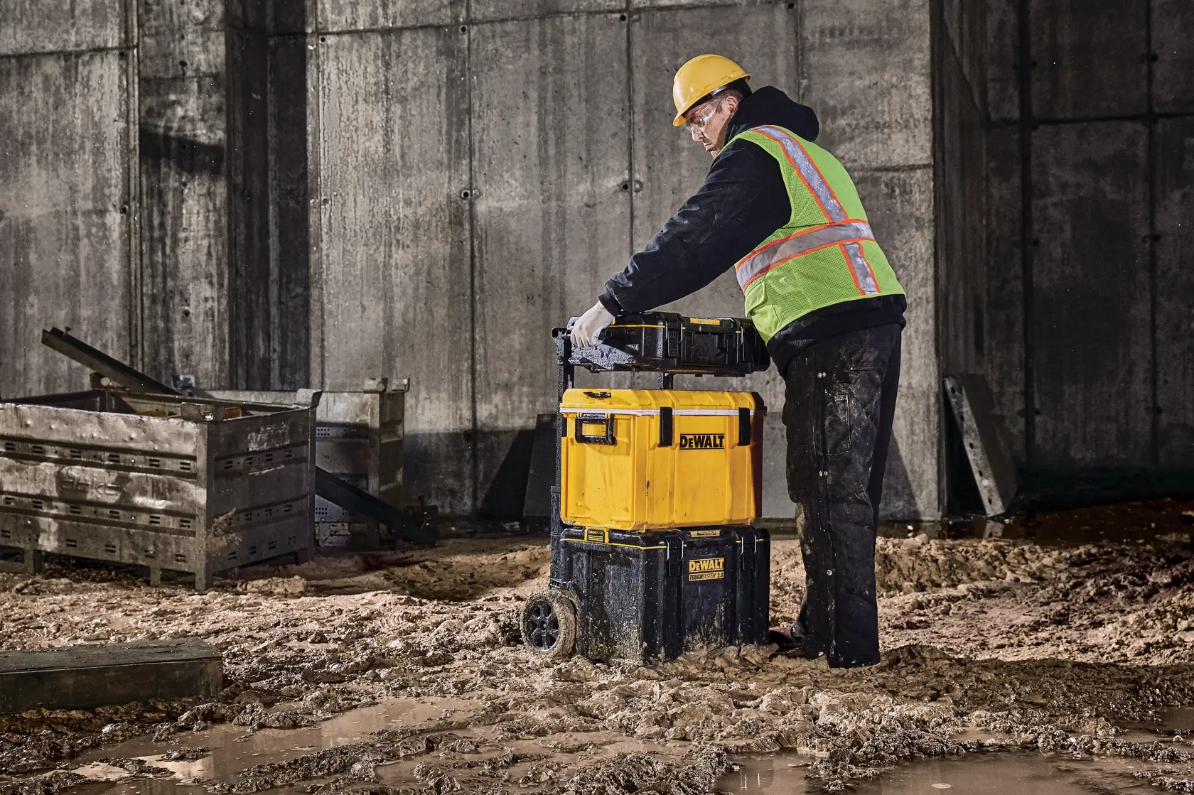 Arbeiter schiebt DEWALT Werkzeugkiste mit Rädern auf schlammiger Baustelle neben Holzkisten.