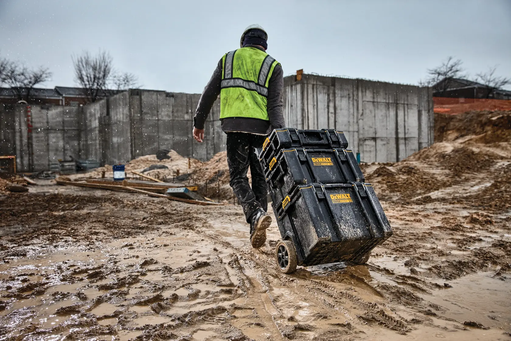 Arbeiter zieht DEWALT Werkzeugkisten auf Rädern über schlammige Baustelle, mit Schutzweste.