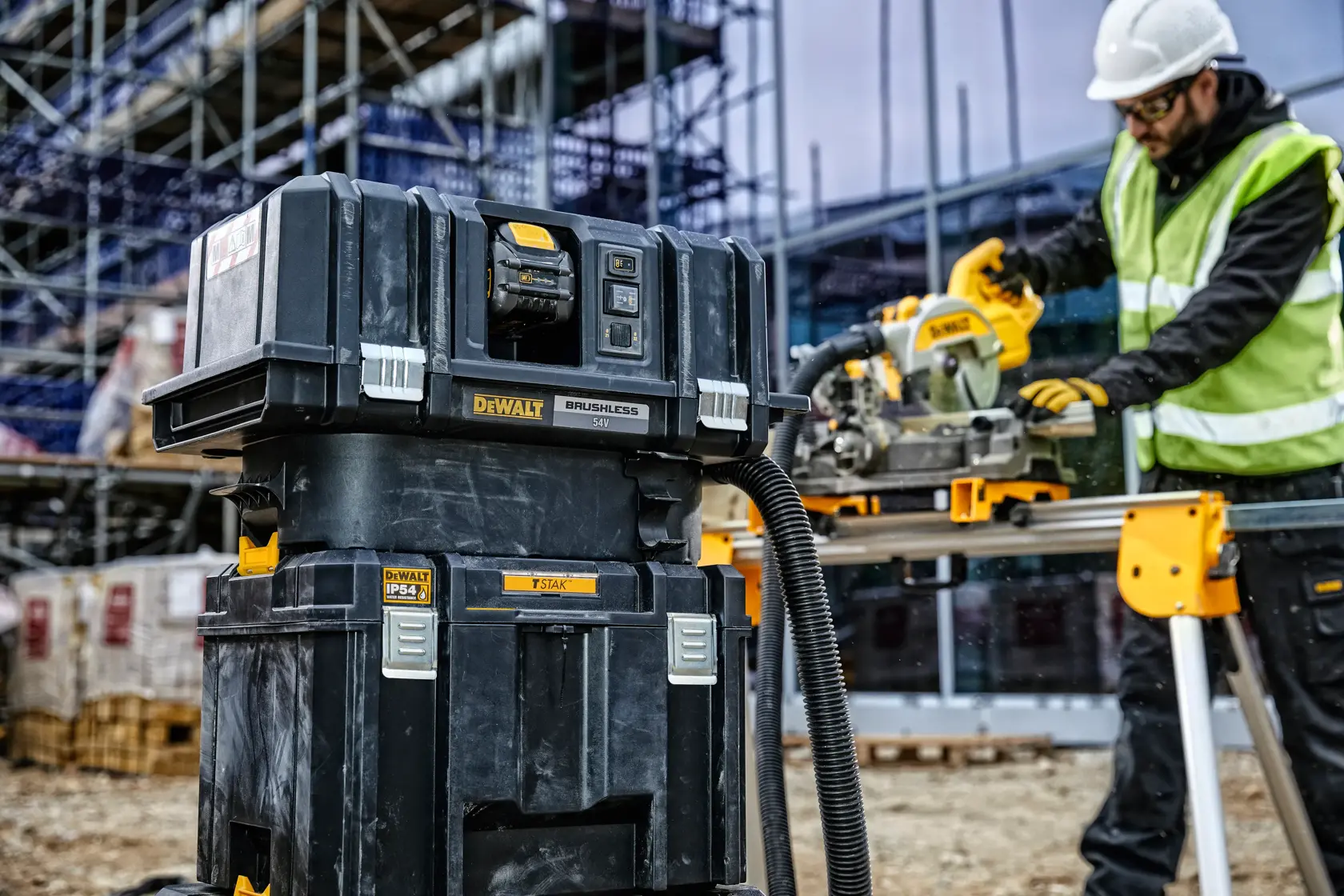 Almacenaje y aspirador DEWALT en obra, trabajador utiliza herramienta eléctrica al fondo.