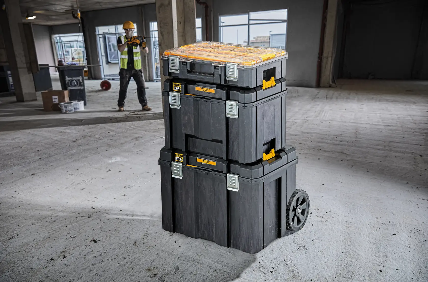 Cajas de herramientas DEWALT apiladas con ruedas en obra, trabajador al fondo.