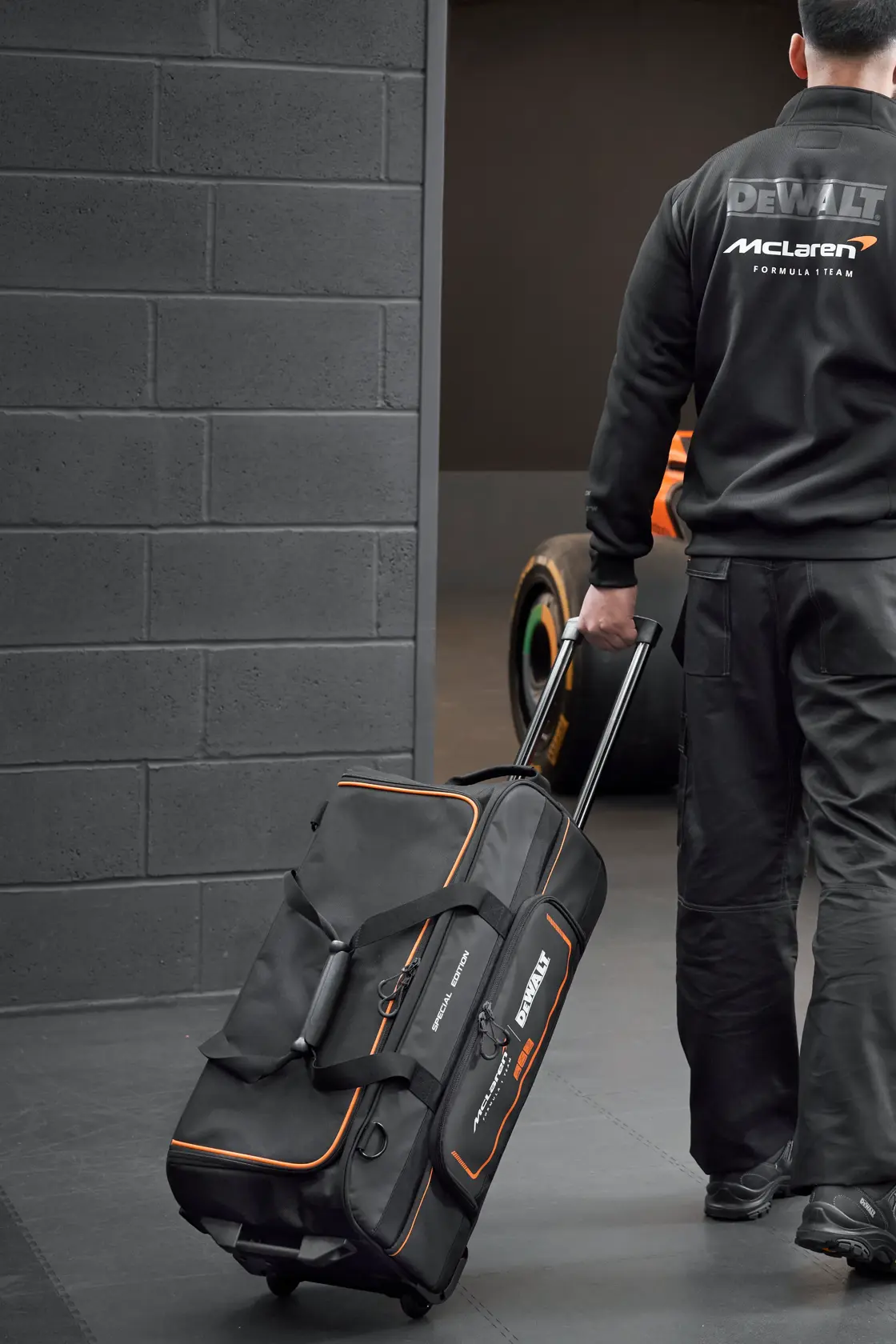 Persona arrastra una bolsa de herramientas DEWALT McLaren con ruedas en un garaje con auto de carreras atrás.
